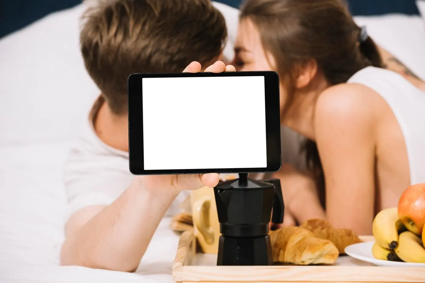 Пара на кровати с едой держит планшет с пустым экраном.